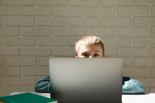 Schoolboy Is Sitting At The Office Desk And Peeking Out From Behind A Gray Laptop Near The Window