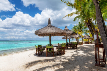 Beach cafe on sandy beach under straw umbrellas, palm trees and beautiful sea on exotic tropical island.