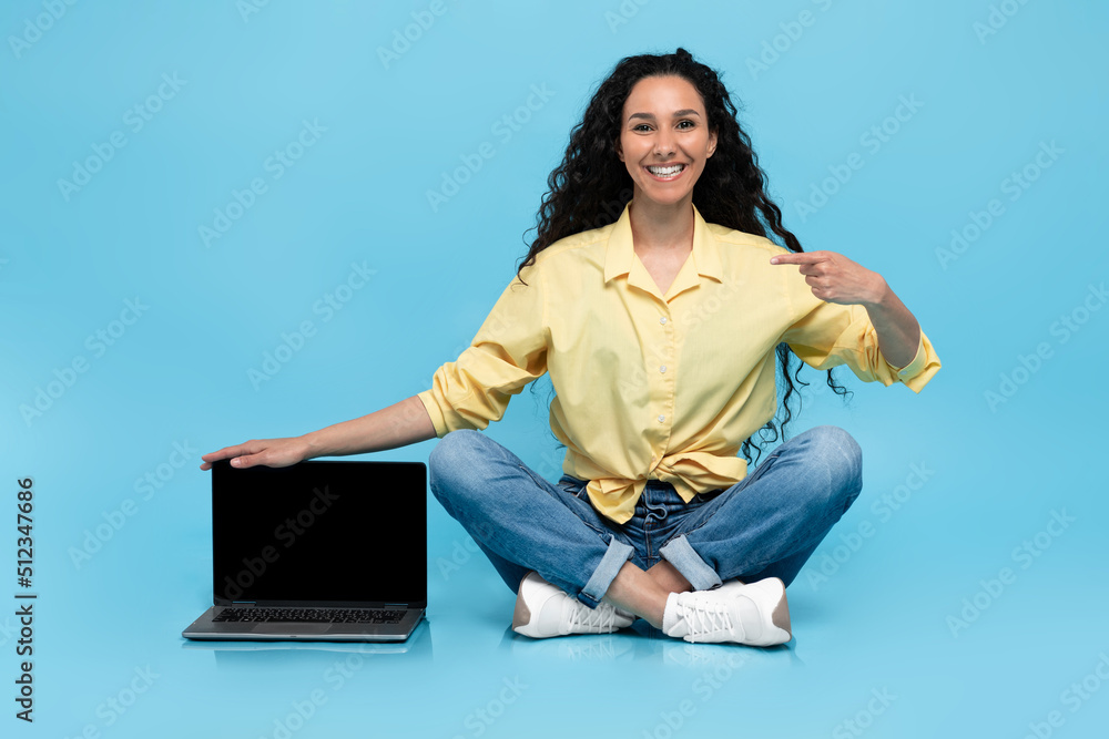Poster young woman pointing at laptop computer with empty screen, offering mockup for website on blue studi