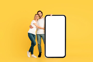 Cheerful millennial caucasian man and female in white t-shirts hugging, stand near huge phone with blank screen