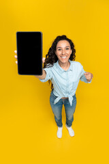 Young woman pointing at tablet with empty screen, offering mockup for website or application over yellow background