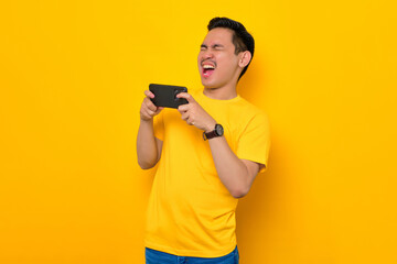 Fototapeta na wymiar Excited young Asian man in casual t-shirt playing online game on mobile phone isolated on yellow background. People lifestyle concept