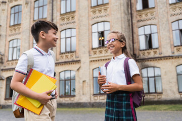 Cheerful interracial pupils with notebooks talking near blurred school outdoors.