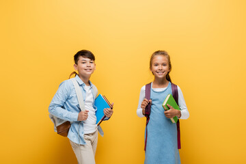 Smiling interracial schoolkids holding books and backpacks on yellow background.