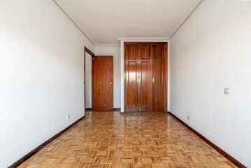 Empty room with shiny oak parquet flooring, plain white painted walls and reddish woodwork on doors...