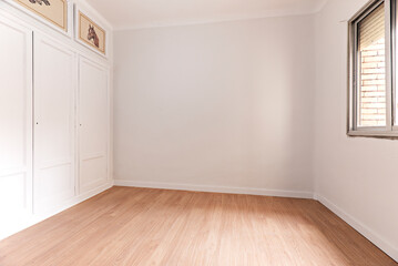 Empty room with oak parquet floor, white painted walls, built-in wardrobe with wooden doors and aluminum window