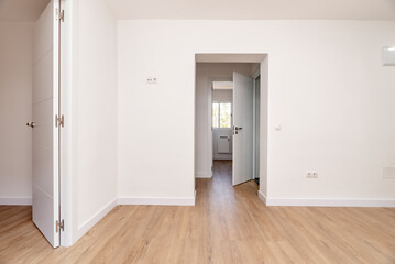 Empty living room with plain white painted walls, several access doors to other rooms with aluminum windows, white carpentry, doors and lamps and light oak floor