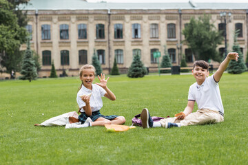 Fototapeta na wymiar Positive schoolgirl holding sandwich and waving hand at camera near asian friend on lawn in park.