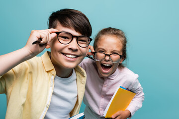 Cheerful multiethnic schoolkids in eyeglasses holding pens and notebooks isolated on blue.