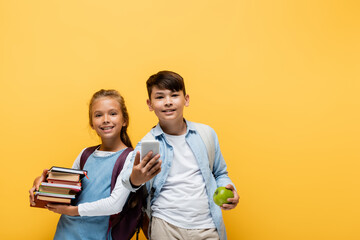 Cheerful interracial schoolkids holding books and smartphone isolated on yellow.