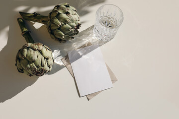 Summer stationery still life scene. Glass of water, green artichoke vegetable in sunlight. Beige table background. Blank paper card, invitation mockup scene with shadows. Flat lay, top view, no people