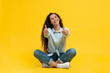 Smiling young woman gesturing thumbs up with both hands, recommending something on yellow studio...