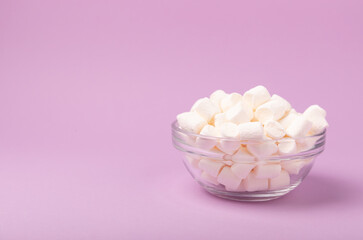 Fototapeta na wymiar Marshmallows on a lilac background in a glass bowl. White marshmallow flat lay. Sweets and snacks for a snack. Chewing candies close-up. Copy space. Place for text. Winter food concept.