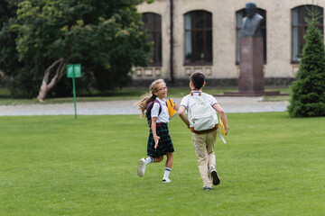 Obraz premium Happy schoolgirl holding notebooks while walking near friend on lawn.