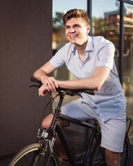 Young modern and happy looking blonde man in short sleeves posing on a bike in city. Stylish guy sitting on bicycle and truly laughing.