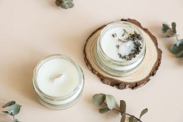 Two candmade candles from paraffin and soy wax in glass with wooden wick and dry herbal isolated on pastel beige background. Flat lay, top view, copy space