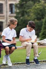 Asian schoolboy writing on notebook near friend on bench outdoors.