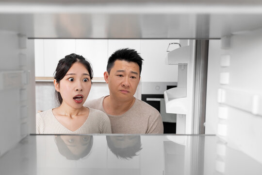 Shocked Asian Couple Looking Inside Empty Fridge At Home