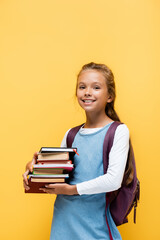 Smiling schoolgirl with backpack holding books isolated on yellow.
