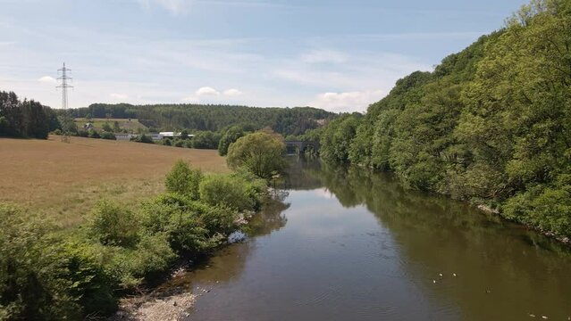 Brown fields and lush deciduous forests growing along the shore of the Sieg river in west Germany. Wide angle aerial view approaching an old train bridge