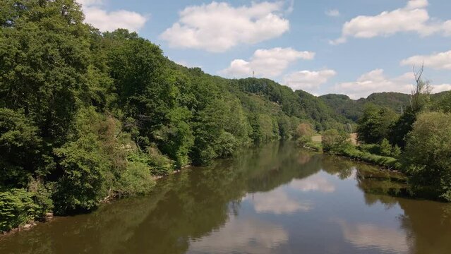 The Sieg flowing calmly in between lush, leafy riverbanks underneath a blue sky with cumulus clouds. Wide angle pull back shot