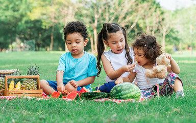 Naklejka na ściany i meble Three little mixed race kids consist of African and caucasian boys and girls smiling with happiness, fun amusement, playing, sitting for picnic and eating piece of watermelon fruit in outdoor garden.