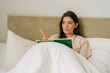 Young brunette female student learning and reading a book in her bed in the morning