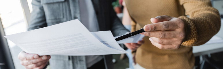 partial view of man pointing with pen at papers near colleague in advertising agency, banner.