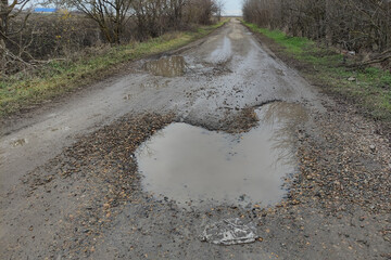 Country road with big puddles through agricultural fields in cloudy weather