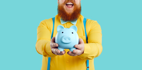 Piggy bank in form of blue pig in hands of joyful contented unrecognizable bearded fat man. Man in...