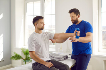 Obraz premium Professional physiotherapist controls his patient who is doing arm muscle rehabilitation exercises. Serious young man doing physical exercises with dumbbells at modern physiotherapy clinic or center