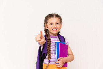 A little girl with a backpack and books points her index finger forward on a white isolated background.