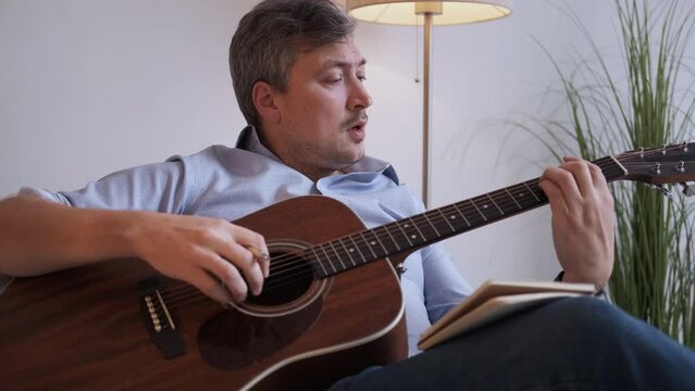 New melody. Inspired man. Composing music. Casual middle-aged guy sitting sofa playing guitar making notes of chords in light room interior.