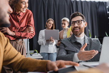 happy advertising manager in eyeglasses pointing at blurred laptop near interracial colleagues.