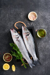 Sea Bass raw. Fresh sea fish bass with salt, pepper, parsley, olive oil and lemon on cutting board on dark concrete rustic background. Food cooking background. Top view, copy space.