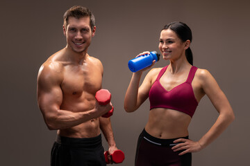 Fototapeta na wymiar Smiling athletic man holding red dumbbell in hand and looking at the camera, while woman drinking water nearby. Indoor studio shot isolated on brown background