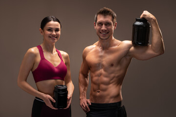 Waist up portrait view of the muscular male and female bodybuilders holding two packs of protein powder and smiling isolated on brown background