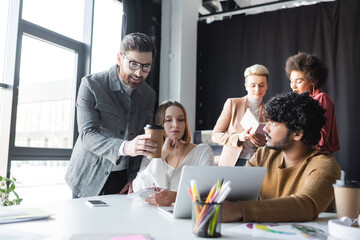 smiling man with paper cup pointing at laptop near interracial advertising managers.