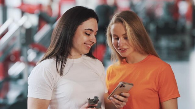 two young beautiful sporty women spend time in smartphone on the background of the gym