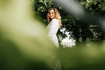 Portrait of smiling young woman wearing delicate white dress with polka dots and standing in frame of foliage and trees. Curly model posing and looking at camera outdoors
