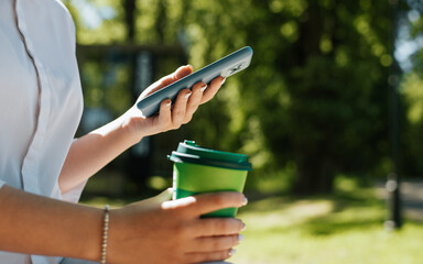 Unrecognizable woman typing on smartphone holding cup of coffee, close-up. Side view of girl using mobile phone, browsing internet on gadget during coffee break outdoors. Selective focus on device