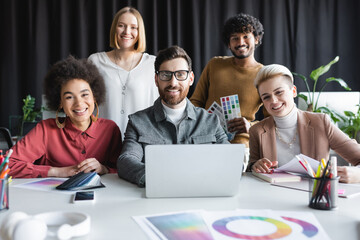 happy multiethnic advertising designers looking at camera near laptop and color samples.