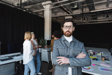 advertising agent in eyeglasses standing with crossed arms near blurred multiethnic colleagues.