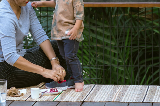 Loving mother helping her little son to put on sneaker while spending time together on balcony at riverside, outdoor on summer day. Child in pretty jeans and gray sport shoes. Friendly happy family.