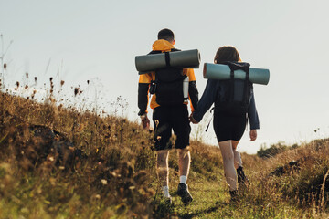 Back View of Hiking Couple, Young Travelers Man and Woman with Backpacks and Camping Mat Holding by Hands and Moving Forward on the Route