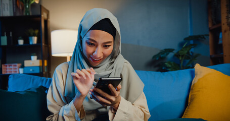 Attractive cheerful young Asian muslim beauty woman in hijab with casual sitting on couch use phone enjoy online shopping on cozy living room in home at night. Girl Islam quarantine lifestyle concept.