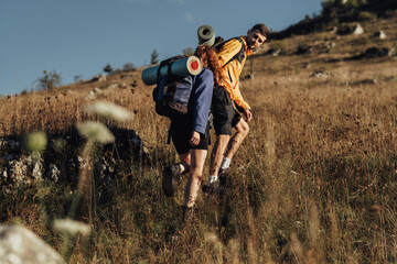 Two Travelers with Backpacks and Camping Mat on a Route, Man Helping Woman to Move Forward on the Top of Hill During Sunset © Romvy