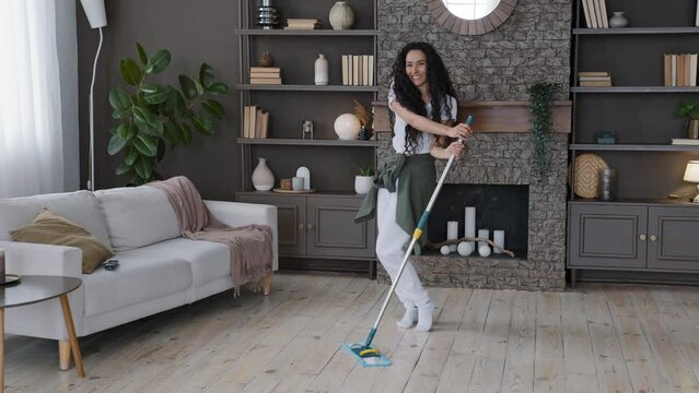 Young cheerful woman cleaning apartment washes floor in living room excited energetic girl dancing with mop comic motion fun housekeeping active moving to music cleans on weekend dancer female at home