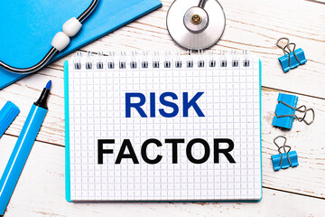 On a light wooden background, a stethoscope, a blue notepad, blue paper clips, a blue marker and a sheet of paper with the text RISK FACTOR. Medical concept