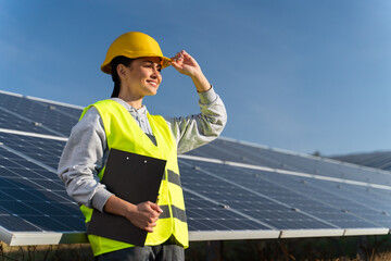 Professional woman engineer looking away with pleasure smile while working at ecological green field with solar panels. Photovoltaic power station concept